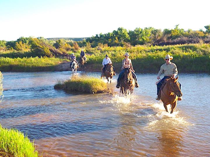 People riding horses through river with grass islandand banks